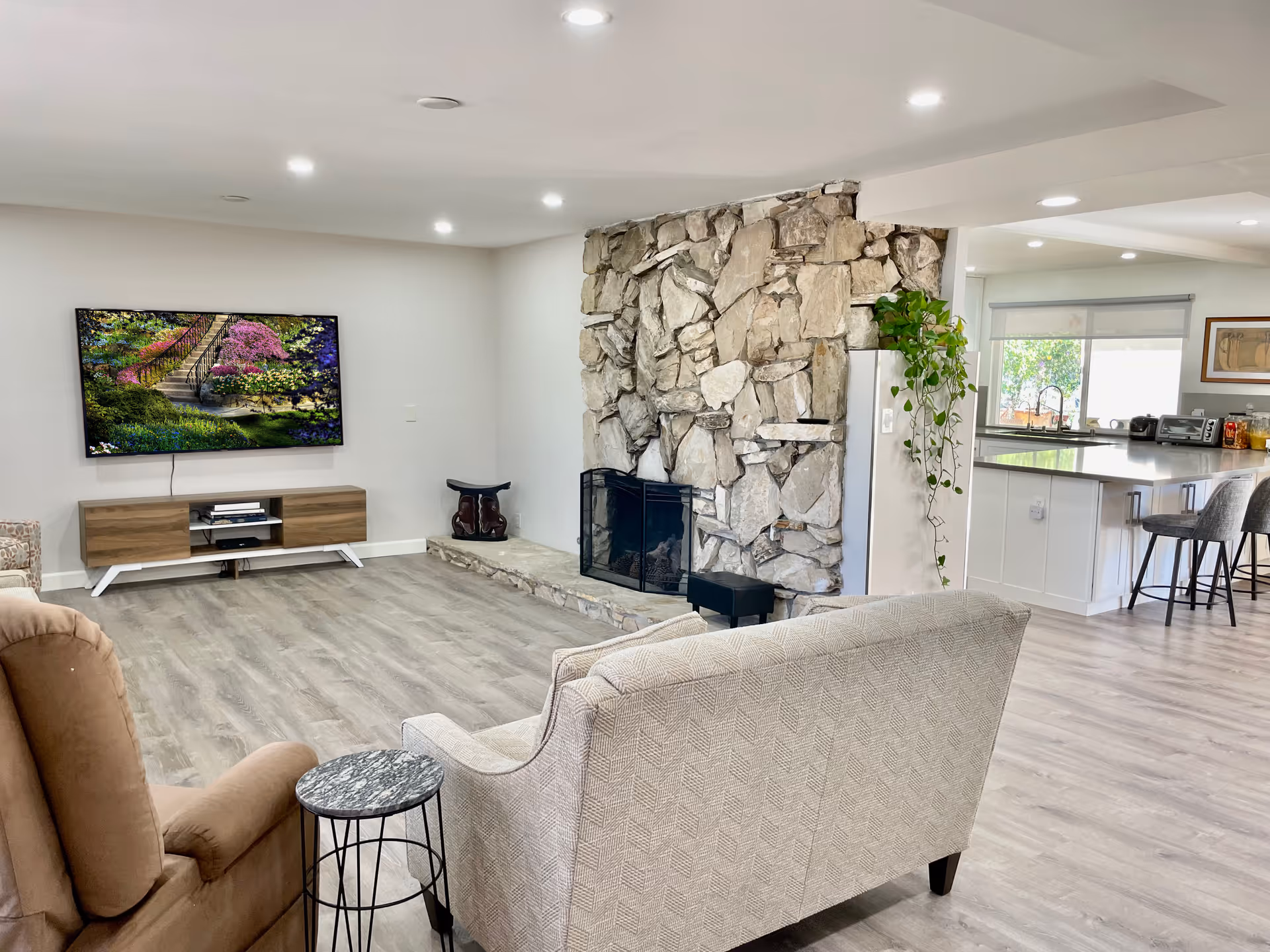 A bright and spacious living room with light wood flooring, a stone fireplace, a wall-mounted flat-screen TV displaying a garden scene, a beige patterned sofa, a brown recliner chair, a small round side table, and an open view into a modern kitchen with white cabinetry and bar stools.