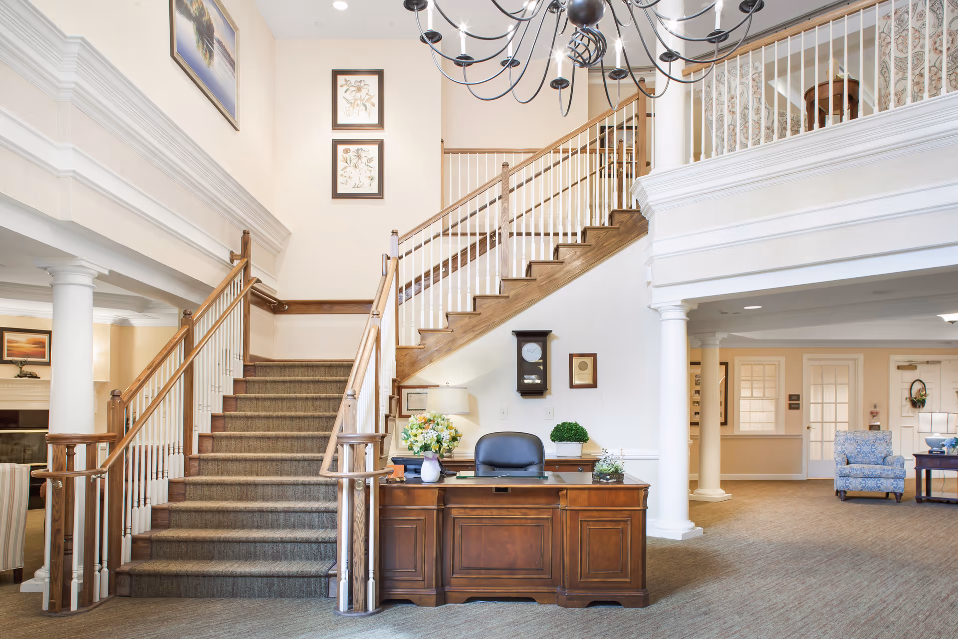 Spacious senior living lobby with a wooden reception desk, carpeted staircase and seating area under a chandelier.