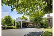 Entrance area of a senior living facility named Spring House Estates, featuring a covered drop-off zone with a white canopy supported by columns, surrounded by green trees and shrubs under a partly cloudy sky.