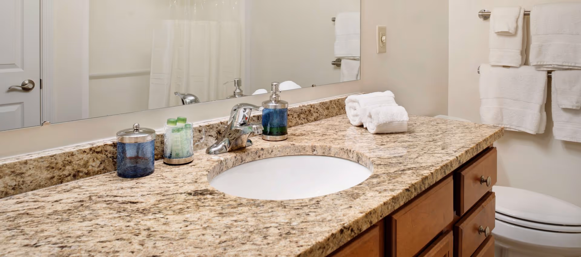 Bathroom countertop with a granite surface, an oval sink, a silver faucet, a soap dispenser, a container with toiletries, and neatly folded white towels. A large mirror is mounted on the wall above the countertop, and a toilet with a towel rack holding white towels is visible in the background.