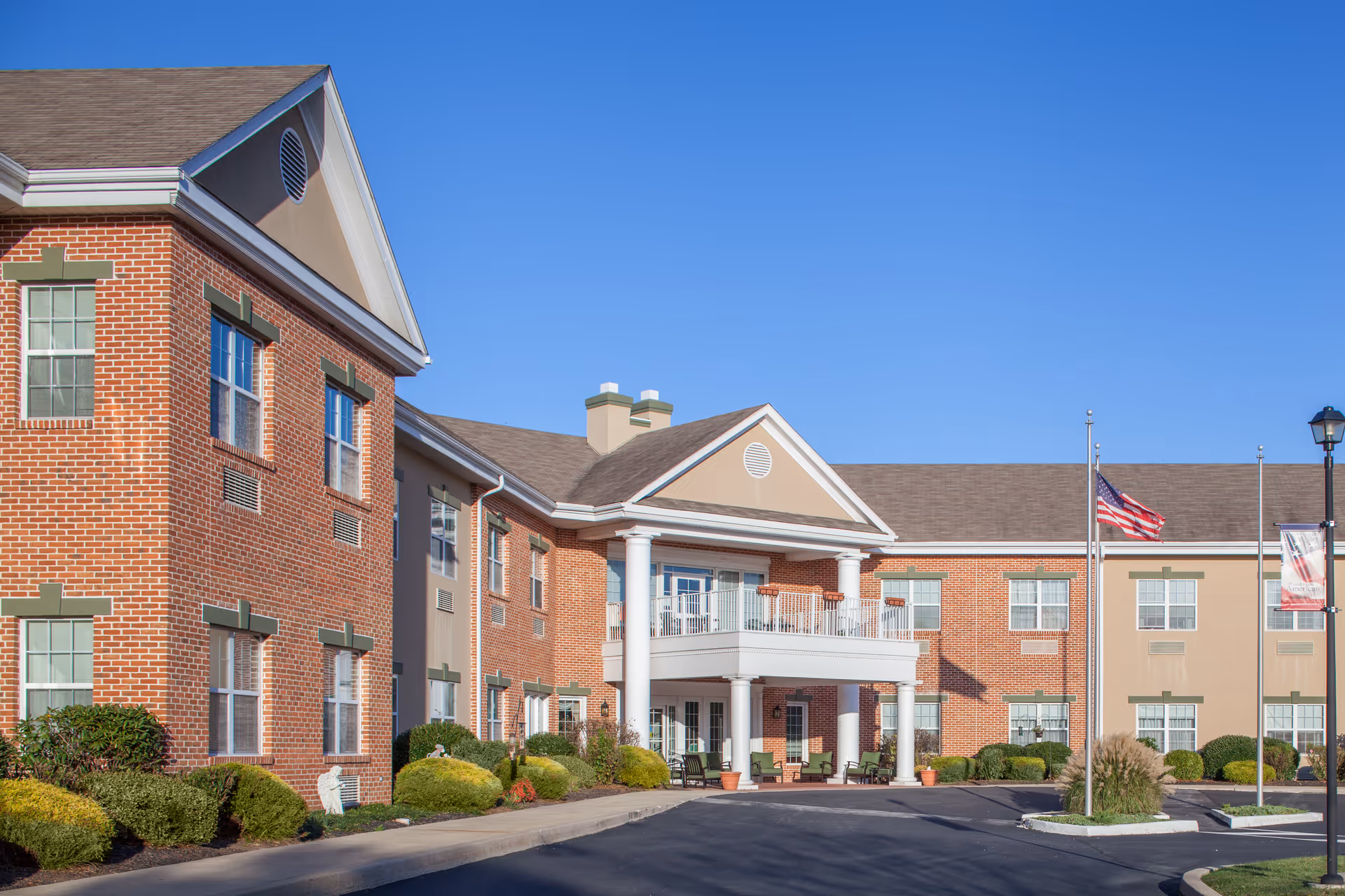 Front entrance of a two-story brick senior living building with a portico, balcony and an American flag under a clear blue sky.