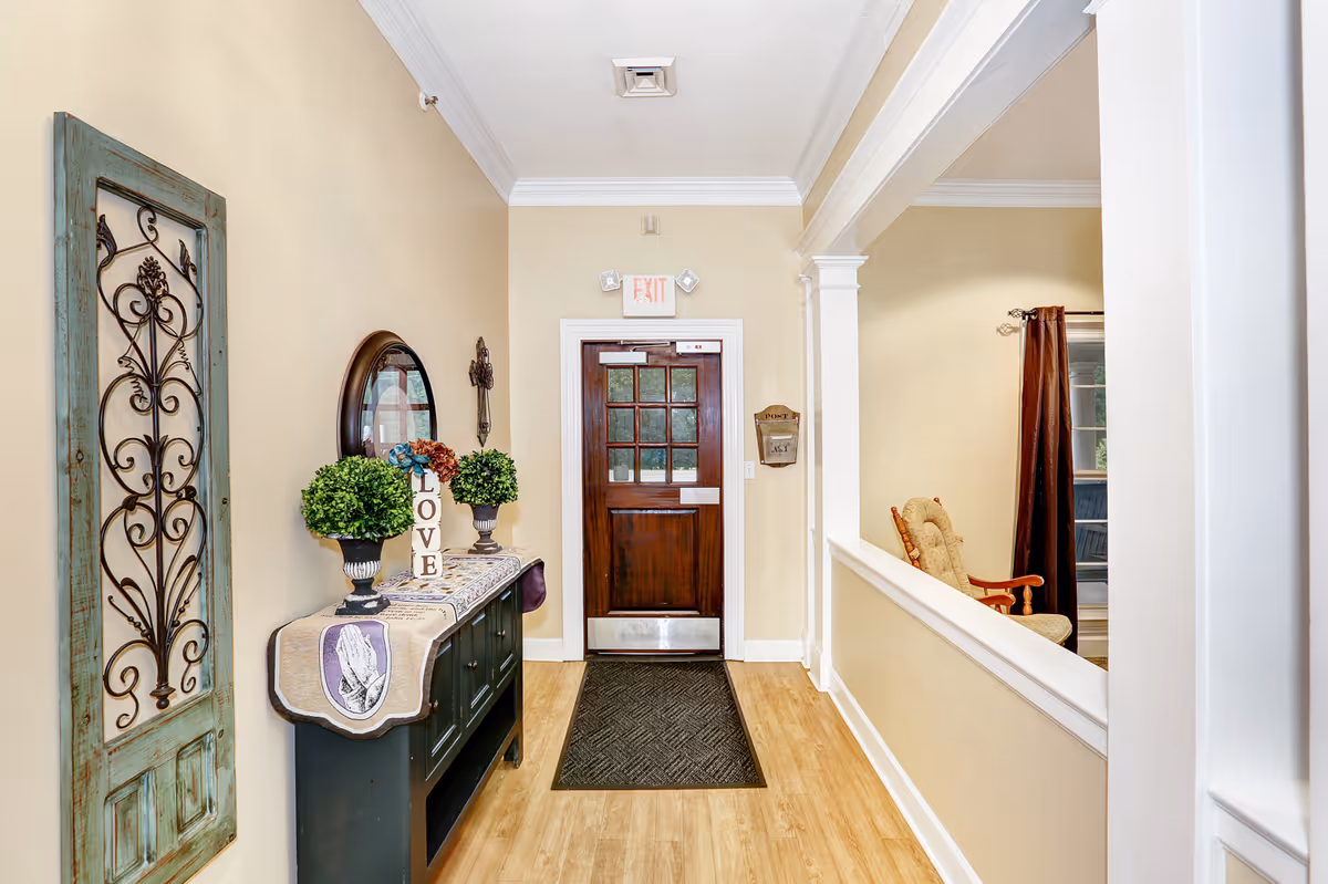 Interior hallway of a senior living facility with a wooden door at the end, a black console table with decorative plants and a 'LOVE' sign, a round mirror on the wall, and a cushioned rocking chair visible through an open partition on the right.