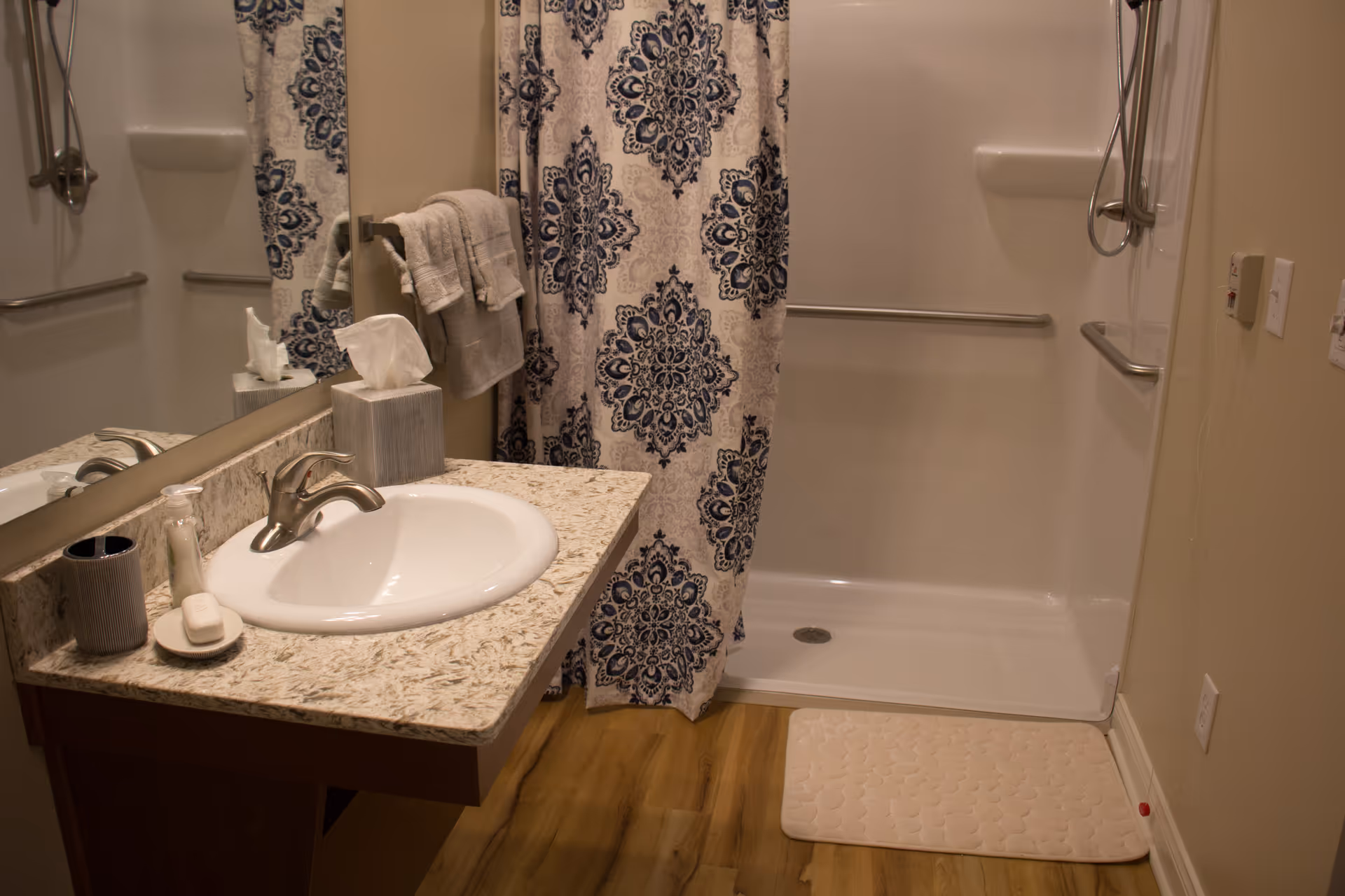 A bathroom with a sink on a granite countertop, a mirror above the sink, a soap dish with a bar of soap, a toothbrush holder, and a tissue box. There is a shower area with a white base and a blue and white patterned shower curtain. The bathroom has wooden flooring and a beige wall with grab bars installed near the shower.