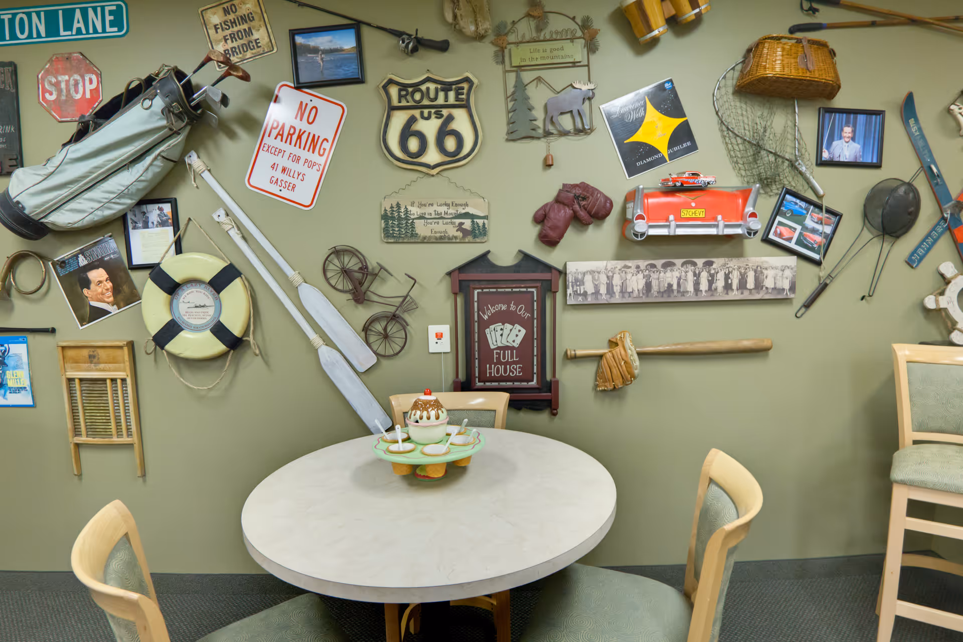 A round table with four chairs in front of a green wall decorated with various vintage and nostalgic items including a golf bag, stop sign, Route 66 sign, boxing gloves, a baseball glove and bat, a fishing net, a miniature bicycle, and framed pictures.