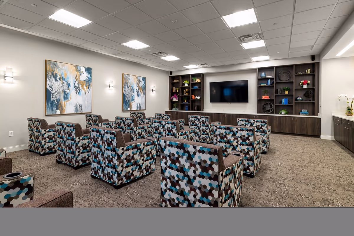 A modern common area with multiple patterned armchairs arranged in rows facing a large flat-screen TV mounted on a wall with built-in shelving. The shelves hold decorative items and plants. The room has a carpeted floor, two abstract paintings on the wall, and bright ceiling lights.