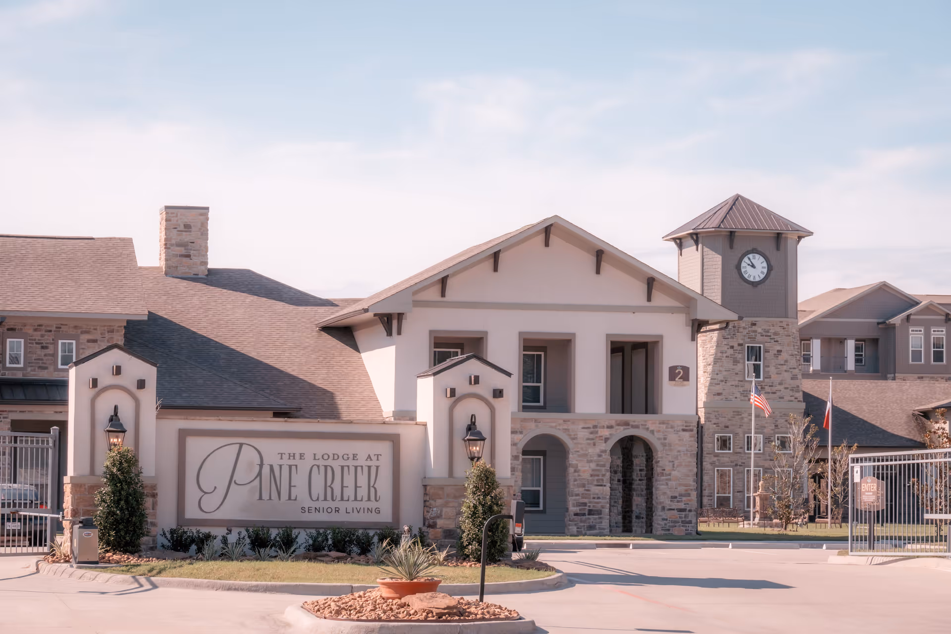 Exterior view of The Lodge at Pine Creek senior living facility featuring a large building with a clock tower, stone and stucco facade, and an entrance sign with landscaping in front.
