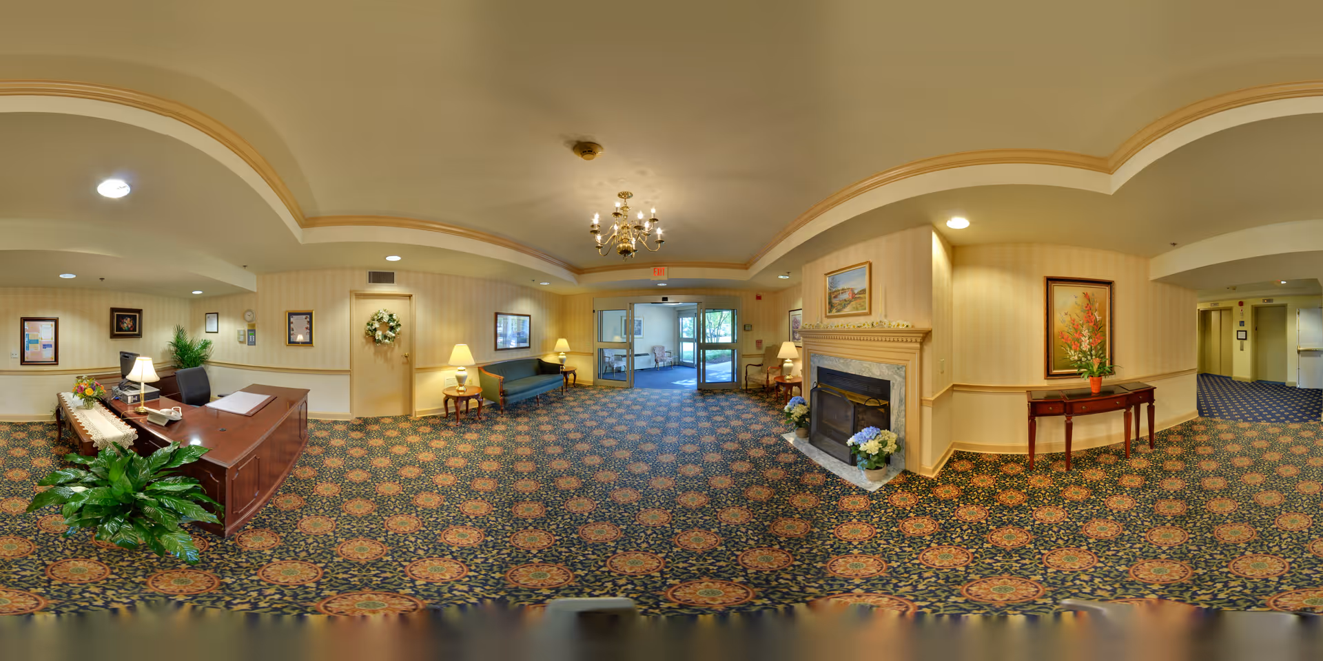 A spacious senior living facility lobby with patterned carpet, a wooden reception desk with a lamp and plant, a seating area with a blue couch and side tables with lamps, a fireplace decorated with flowers and a painting above it, and a table with a flower arrangement. The walls are light-colored with a chair rail, and there are ceiling lights and a chandelier. Glass doors lead outside.