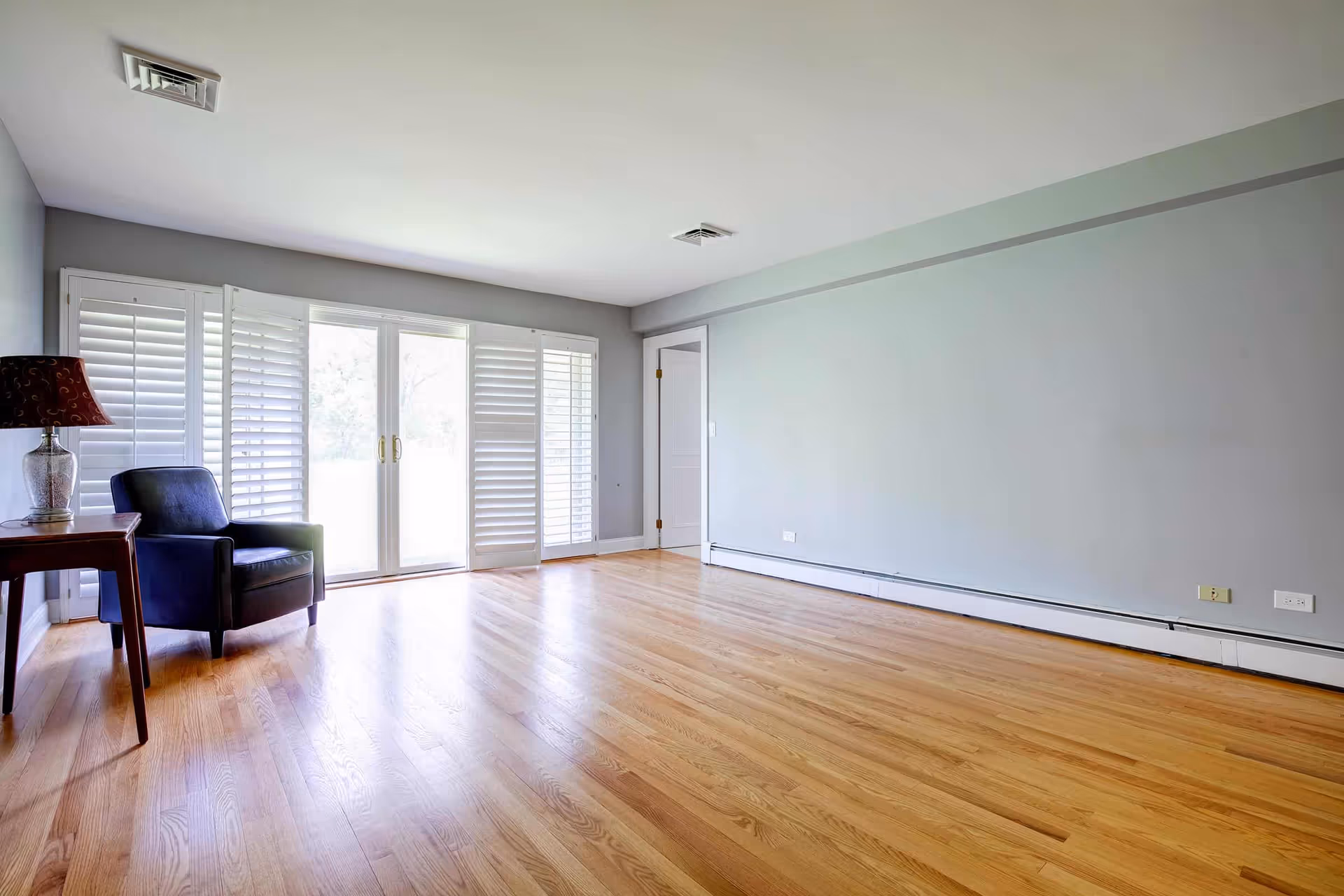 A bright, empty room with light gray walls and polished hardwood floors. There is a single dark blue armchair next to a wooden side table with a decorative lamp on it. Large glass sliding doors with white plantation shutters allow natural light to fill the room. A white door is visible on the right wall.