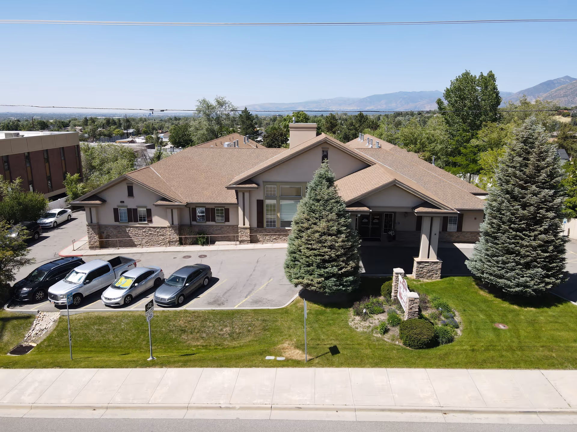 Exterior view of a single-story senior living facility building with a brown roof and beige walls, surrounded by green trees and a well-maintained lawn. Several cars are parked in the parking lot in front of the building. Mountains are visible in the background under a clear blue sky.