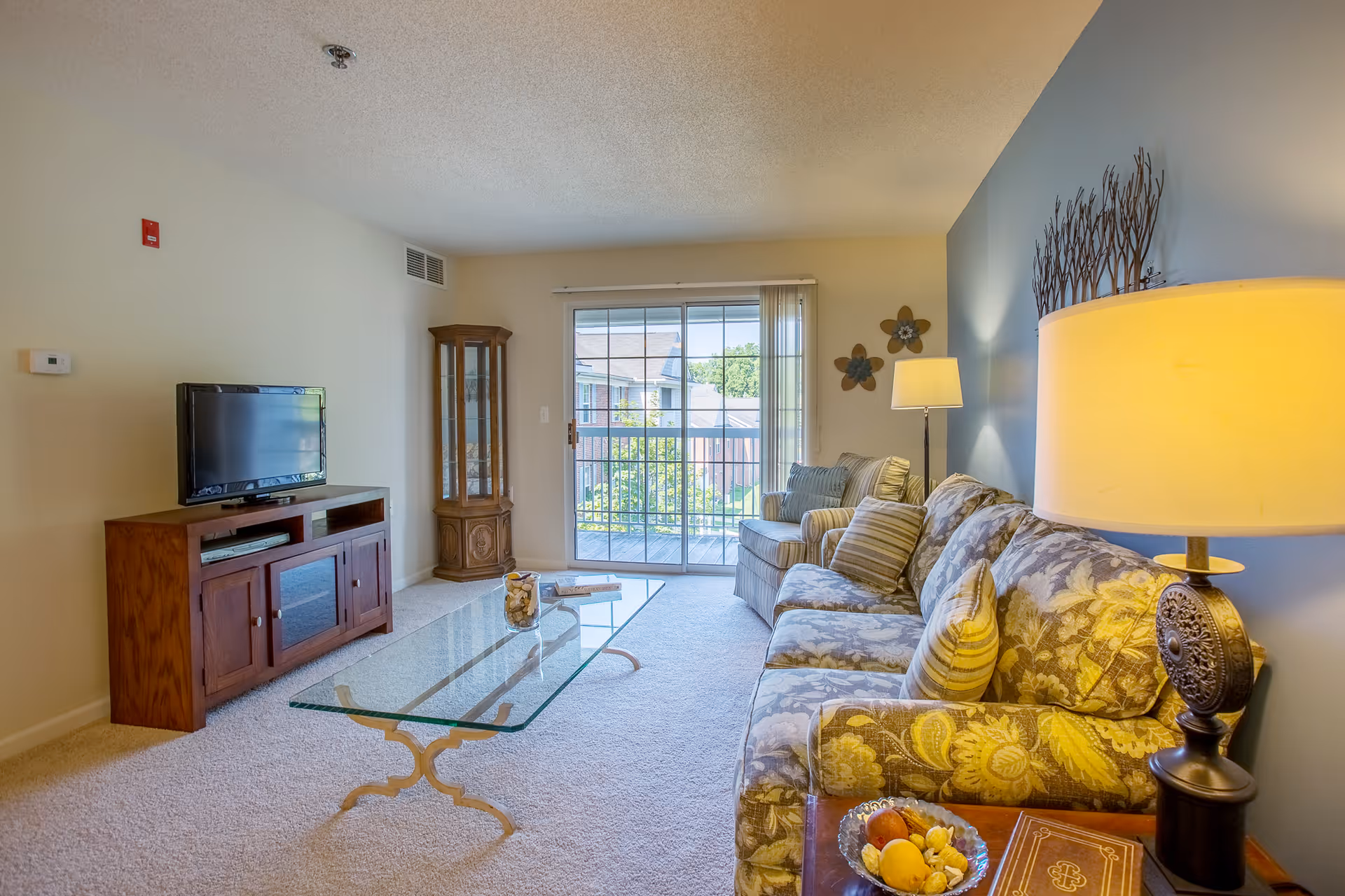 Bright living room with a patterned sofa and armchair, glass coffee table, TV on a wooden console, and a sliding glass door to a balcony.