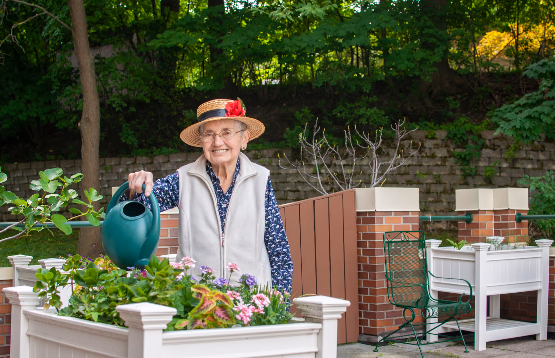 An elderly woman wearing a straw hat with a red flower, glasses, a blue patterned shirt, and a light gray vest is watering plants in a raised garden bed outdoors. She is smiling and standing in a garden area with green trees and a brick wall in the background. There is a green metal chair next to another raised garden bed nearby.