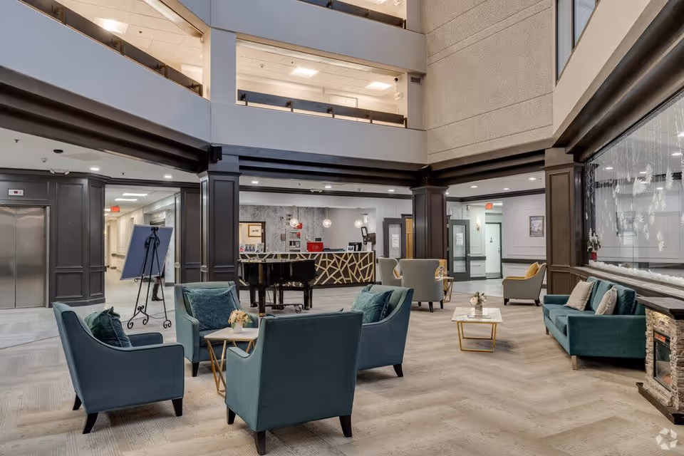 Spacious and well-lit senior living facility lobby with teal armchairs and sofas arranged around small tables, a black grand piano near the reception desk, and multiple floors visible with railings overlooking the area.
