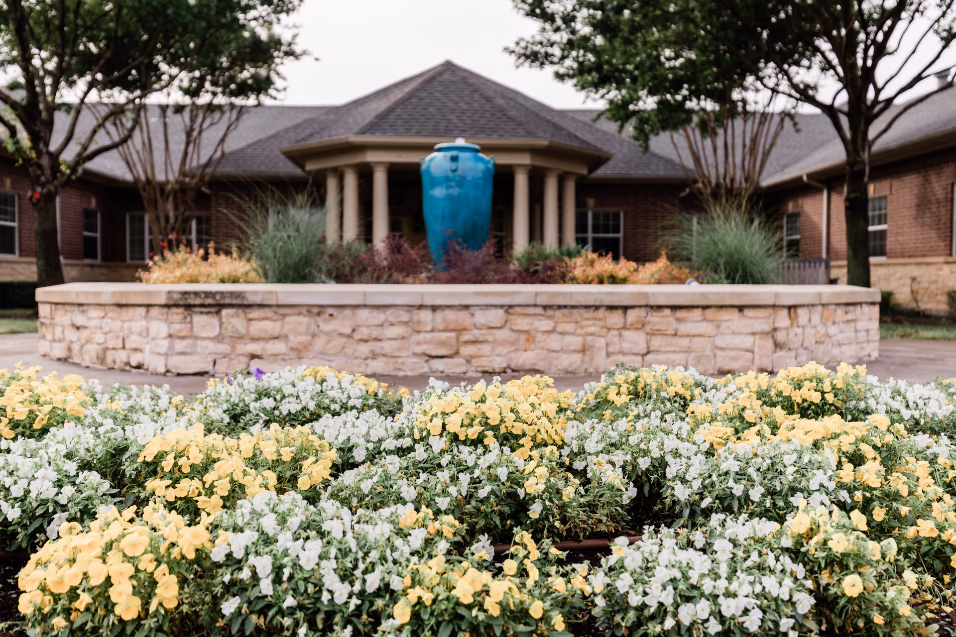 A landscaped outdoor area featuring a stone planter filled with yellow and white flowers in the foreground. Behind the planter is a large blue decorative urn surrounded by plants, with a brick building and trees in the background.