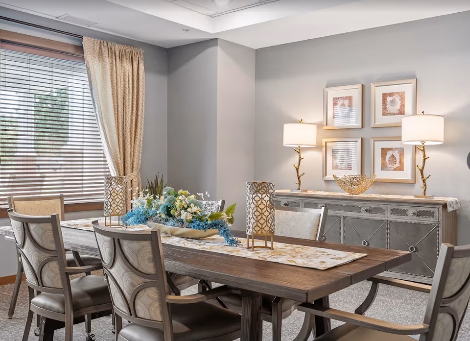 Dining room interior featuring a wooden table and chairs with a floral centerpiece, a sideboard with lamps and framed art, and a window with blinds.