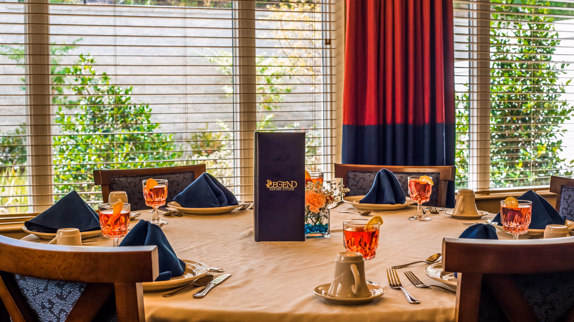 A round dining table set for six with white tablecloth, blue folded napkins, glasses of iced tea with lemon slices, white coffee cups, silverware, and a menu standing upright in the center. The background shows large windows with blinds and greenery outside.