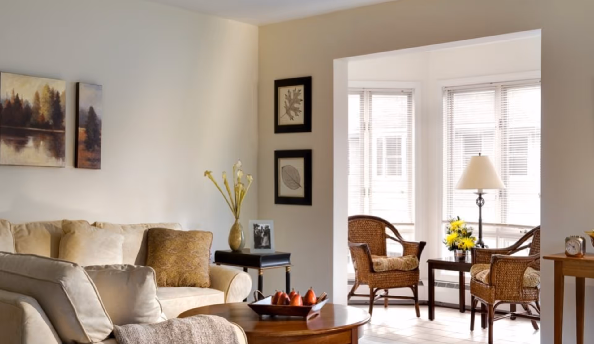 A cozy living room with beige sofas, a wooden coffee table with a decorative bowl of fruit, and a side table with a vase of flowers and a framed photo. In the background, a sunlit sitting area with wicker chairs, a small table with a lamp, and a vase of yellow flowers is visible through a large window.