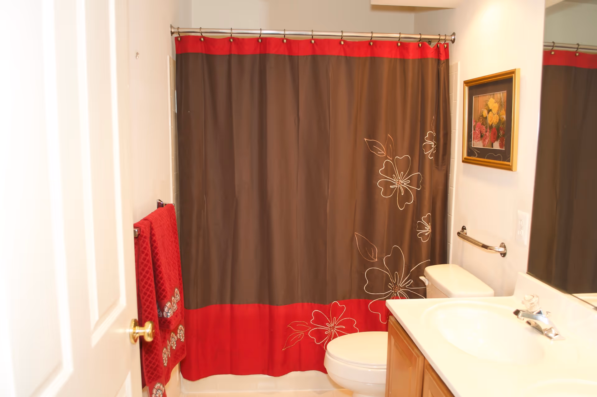 A bathroom with a shower curtain that is brown with a red border and floral designs. There is a white toilet, a sink with a countertop, a mirror, a towel rack with red towels, and a framed floral picture on the wall.