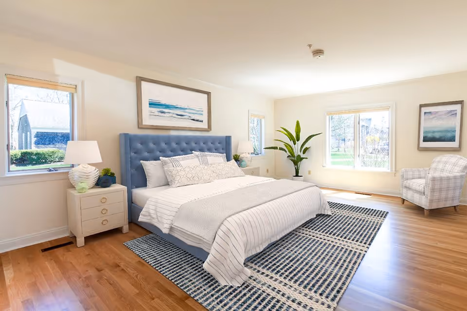A bright and spacious bedroom with a large bed featuring a blue tufted headboard, white and patterned bedding, and a blue and white checkered rug underneath. There are two windows letting in natural light, a white nightstand with a lamp and small plants, a large green potted plant in the corner, and a plaid armchair near the window. The walls are light-colored with framed artwork hanging above the bed and near the armchair.