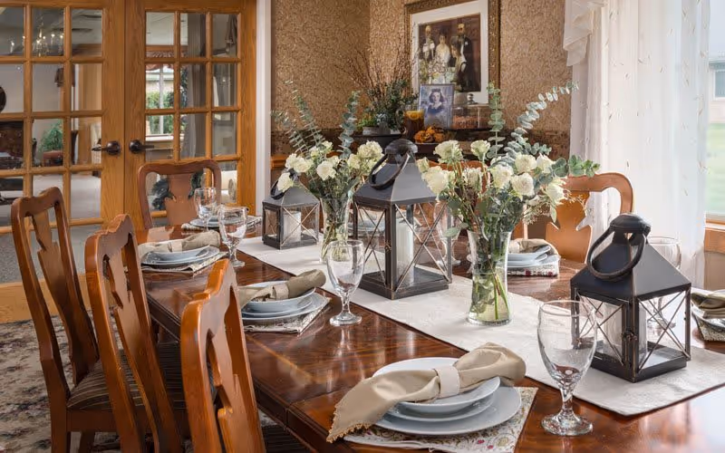 A formal dining room with a polished wooden table set for six. The table is decorated with three black lanterns and three vases filled with white flowers and greenery. Each place setting includes a plate, a folded beige napkin, and a clear wine glass. The room has wooden chairs with striped cushions, a patterned rug, and a sideboard with framed pictures and decorative items. French doors with glass panes lead to another room, and sheer white curtains cover the windows.