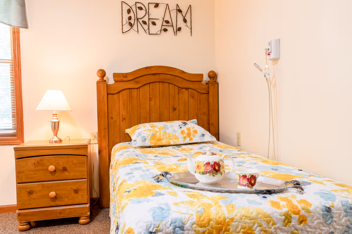 A cozy bedroom with a wooden bed frame and a floral-patterned bedspread in yellow, blue, and white. A matching pillow rests on the bed. On the bed is a silver tray holding a floral teapot and teacup. To the left of the bed is a wooden nightstand with two drawers and a lit table lamp. A window with blinds is partially visible on the left wall. Above the bed, the word 'DREAM' is displayed in decorative wire art on the wall. The walls are painted a light cream color.