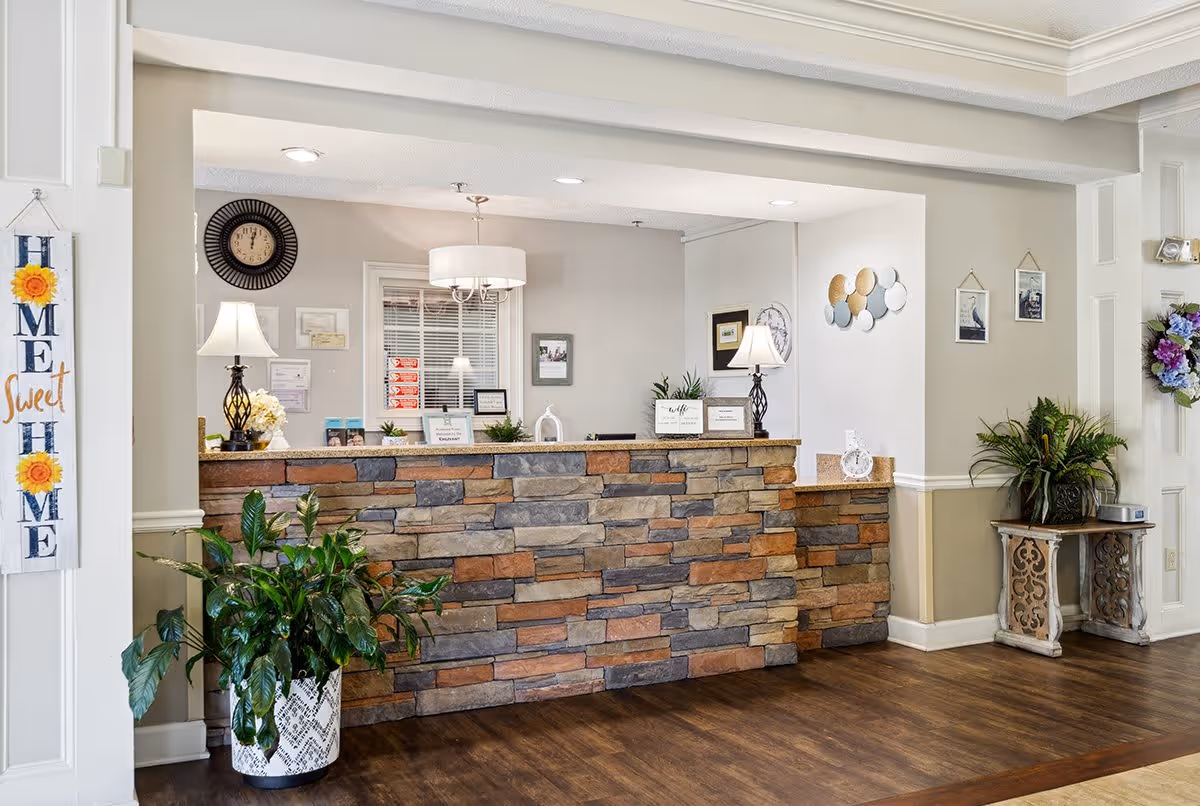 Reception area with a stone-faced front desk, two table lamps, wall clock, decorative plants, and wall art. A 'Home Sweet Home' sign hangs on the left wall, and a small table with a plant and a wreath is on the right side.