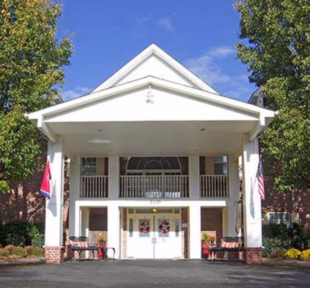 Front entrance of a brick assisted living building with a covered white portico, double doors, flags, and benches.