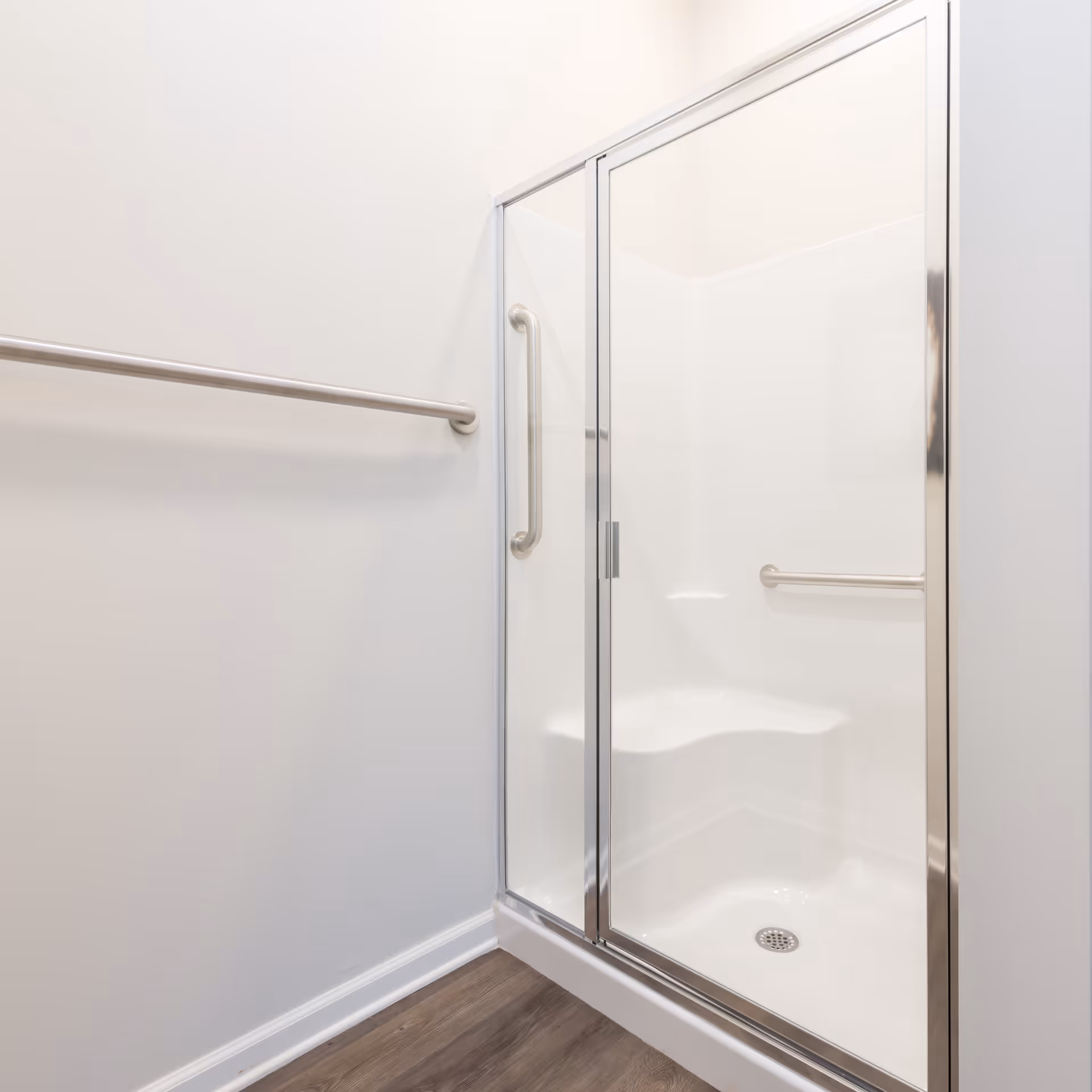 A clean, white shower stall with a glass door and metal frame, featuring a built-in seat and grab bars inside. The surrounding walls are white, and there is a metal towel bar mounted on the adjacent wall. The floor outside the shower is wood-toned.
