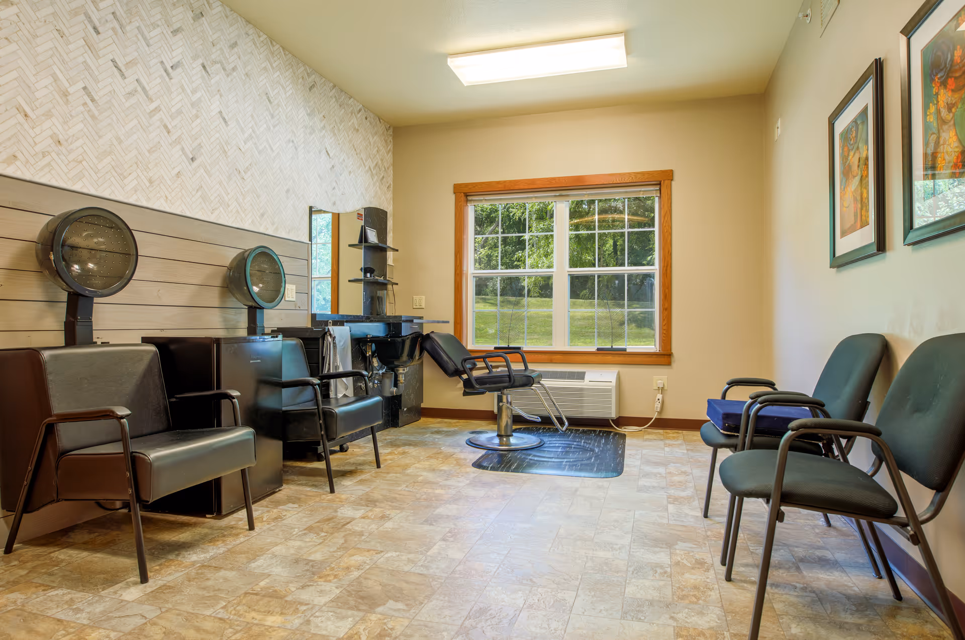 Small salon-style room with styling chairs, hooded hair dryers, a shampoo station, waiting chairs, and a large window.