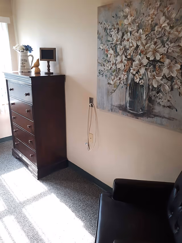 A corner of a room with a dark wooden chest of drawers topped with a white pitcher holding flowers, a small decorative bird, and a small framed item. A large floral painting hangs on the wall above a black leather chair. Sunlight streams through a window, casting shadows on the carpeted floor.