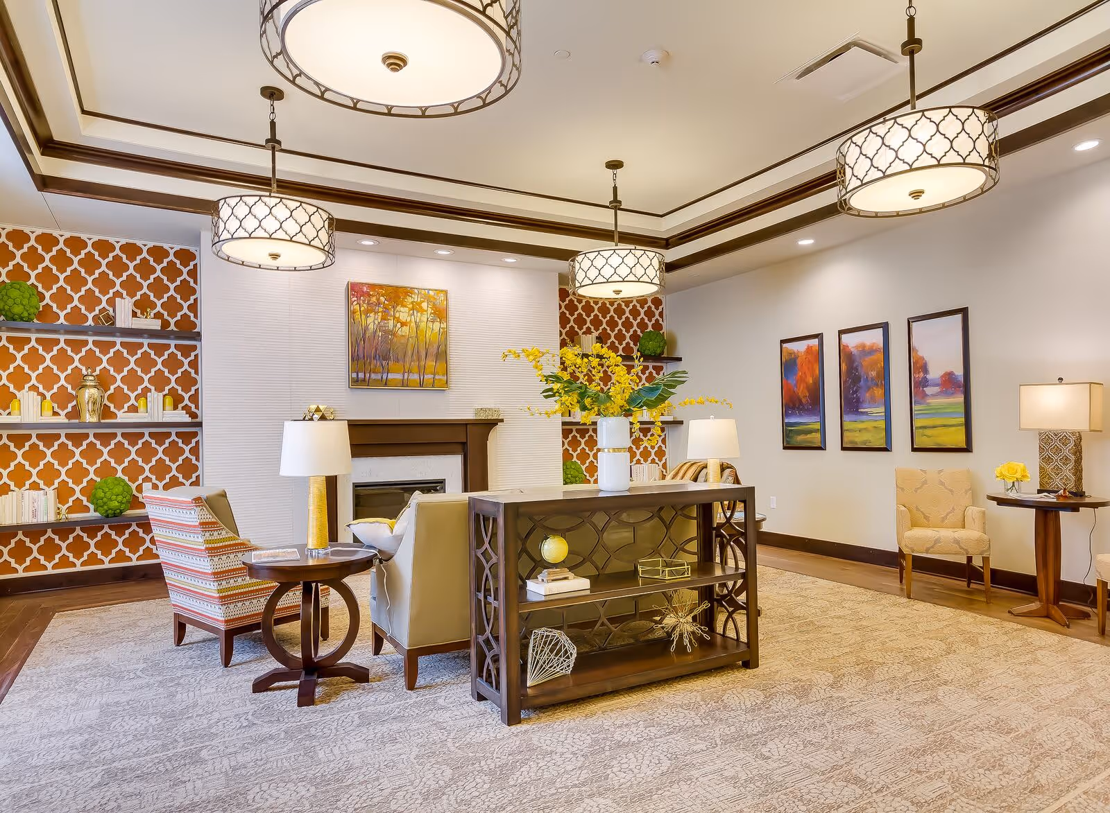 A spacious and well-lit living room with a beige carpet, patterned armchairs, a sofa, and a wooden console table with decorative items. The room features a fireplace with a colorful painting above it, built-in shelves with books and decor, and three large pendant lights hanging from a tray ceiling. The walls are decorated with three landscape paintings and a table lamp with a yellow flower arrangement.