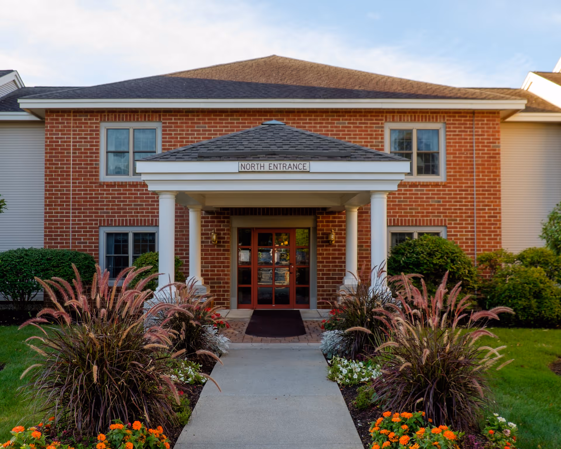 Front view of a brick building with a covered entrance labeled 'North Entrance'. The entrance has white columns and a pathway leading to double glass doors. There are landscaped bushes and flowers on either side of the pathway.