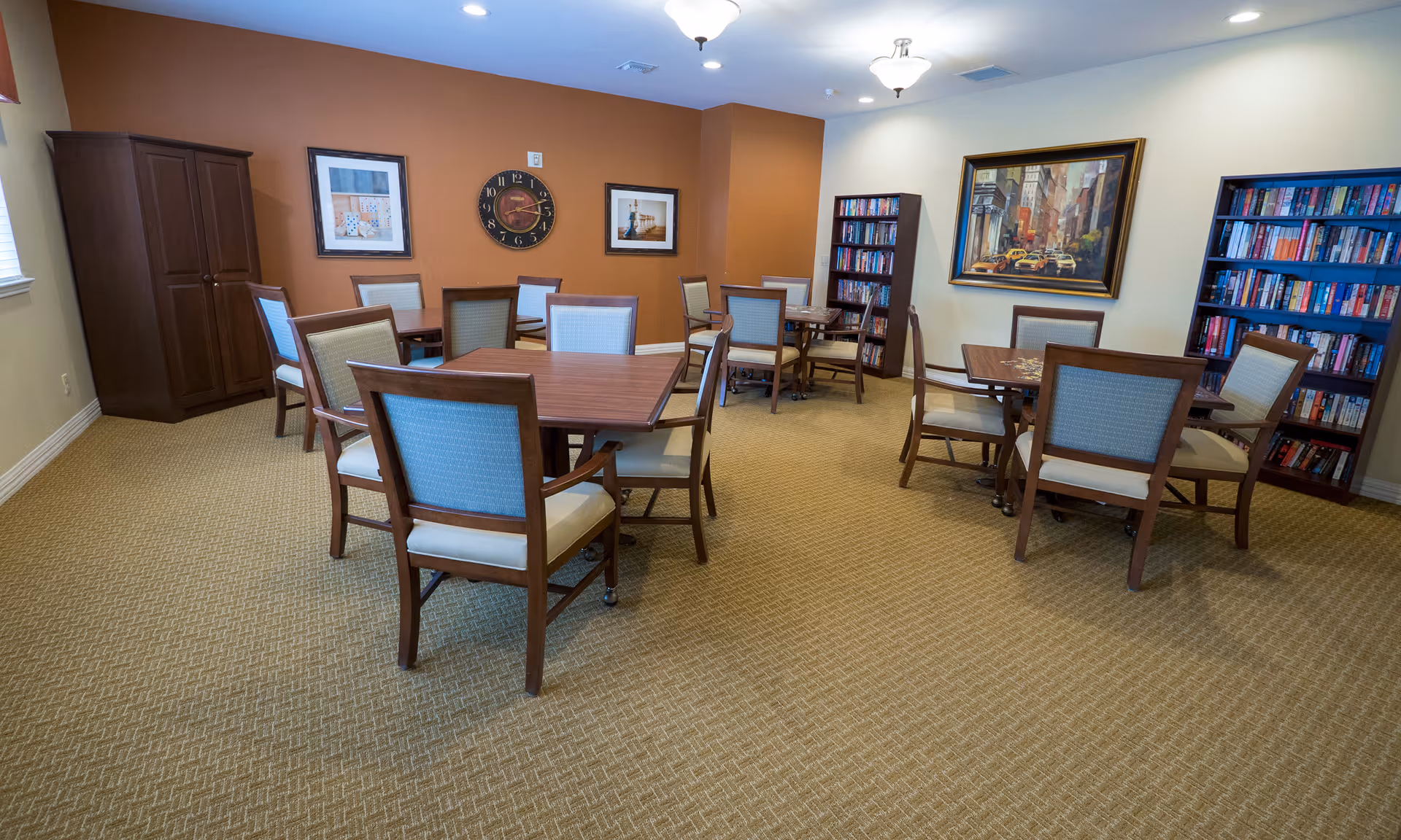A well-lit senior community common room with multiple tables and chairs, bookshelves, and framed artwork on the walls.