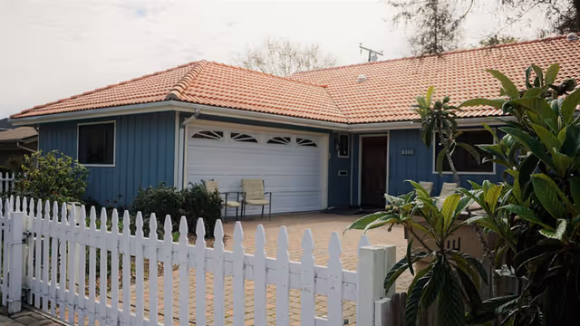 Exterior view of a single-story residential building with a red tiled roof, blue siding, a white garage door, and a white picket fence in the foreground. There are two chairs placed near the garage door and some greenery around the entrance.