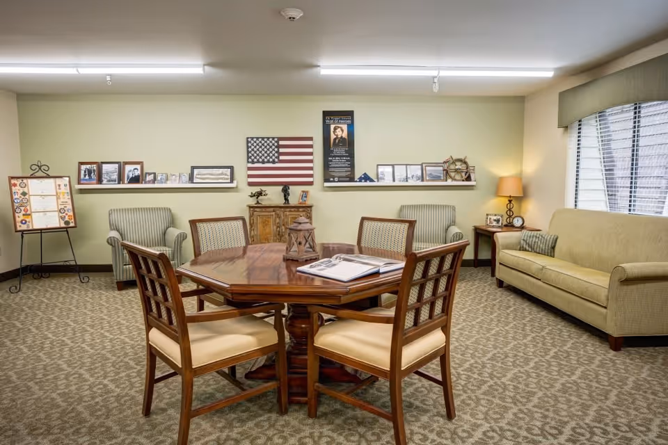 A cozy sitting area in a senior living facility with a wooden octagonal table surrounded by four cushioned chairs in the center. The room features a beige carpet, two striped armchairs, a beige sofa, and a side table with a lamp and clock. The walls are decorated with framed photos, an American flag, and a display board on an easel. A large window with blinds allows natural light into the room.
