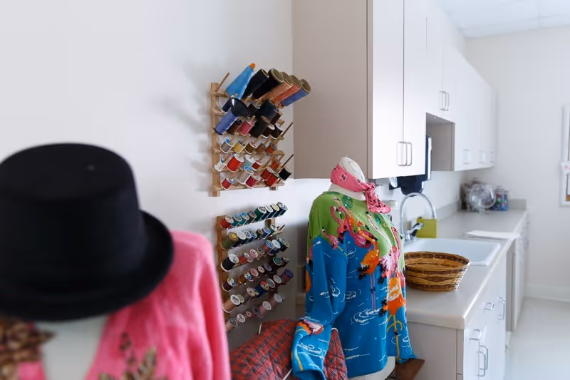 Interior view of a craft or sewing room with two mannequins dressed in colorful clothing, one wearing a black hat and pink jacket, the other in a blue and green sweater with a pink scarf. There are multiple spools of thread organized on wall racks, white cabinets, a countertop with a basket, and a sink in the background.