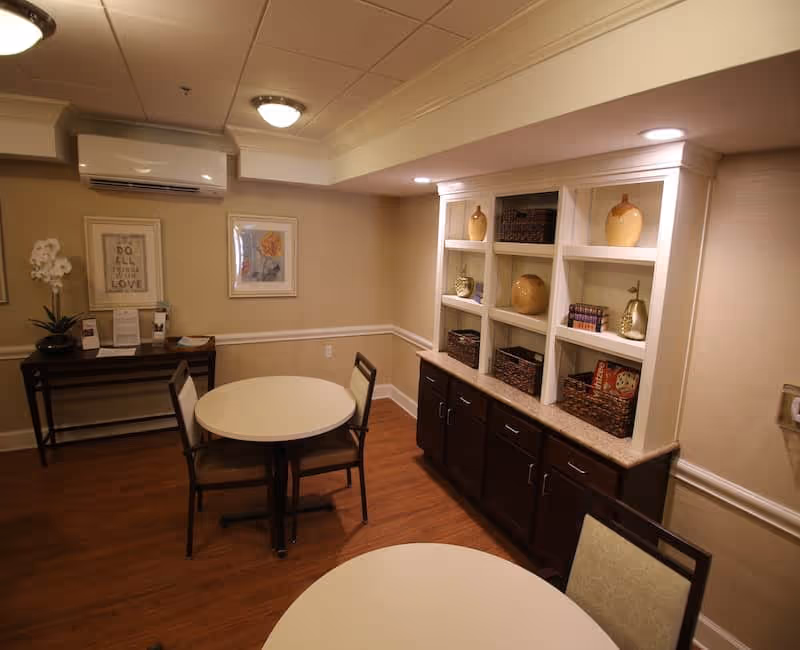 A cozy interior room with two round tables and chairs, wooden flooring, a built-in cabinet with decorative vases and baskets, framed artwork on the walls, and a small console table with a plant and informational materials.