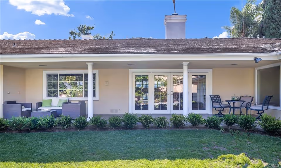 Outdoor patio area of a single-story building with beige walls and a shingled roof. The patio features two seating areas: one with dark wicker furniture and green cushions on the left, and another with a small round table and four metal chairs on the right. There are white pillars supporting the roof over the patio, and a row of green shrubs lines the edge of the patio. The sky is blue with some clouds visible above the roof.