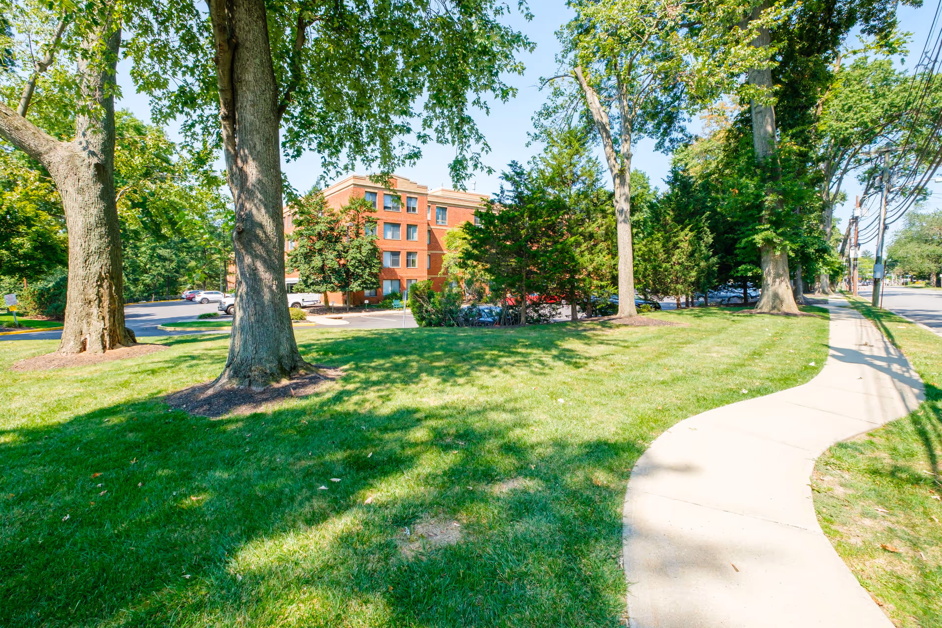 Green lawn with large trees and a curved sidewalk leading toward a multi-story brick residential building set back among trees.