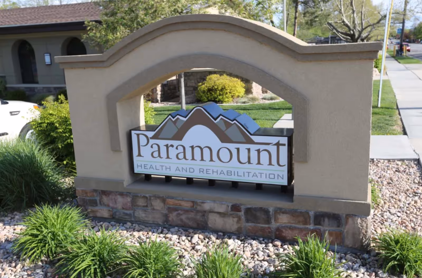 Outdoor stone and stucco sign for Paramount Health and Rehabilitation with a mountain logo, surrounded by landscaping and a sidewalk nearby.