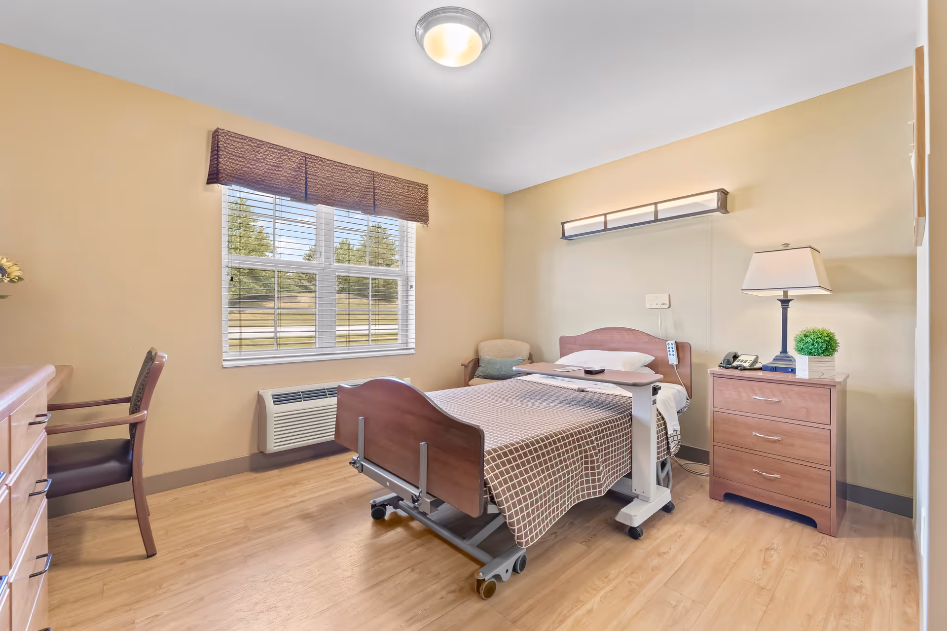 A bright and clean nursing facility bedroom with a hospital bed covered in a checkered blanket, a bedside table with a lamp and a small plant, a chair in the corner, a window with blinds and a valance, and a wooden dresser with a chair next to it.