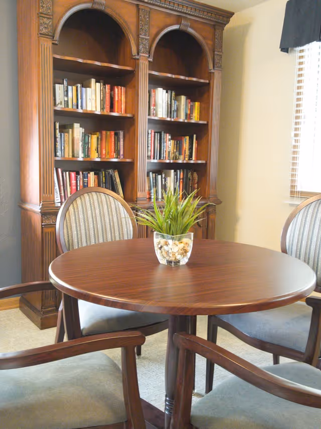 A cozy interior room featuring a round wooden table with a small plant centerpiece, surrounded by four cushioned wooden chairs. Behind the table is a large wooden bookshelf filled with books. A window with blinds is partially visible on the right side.