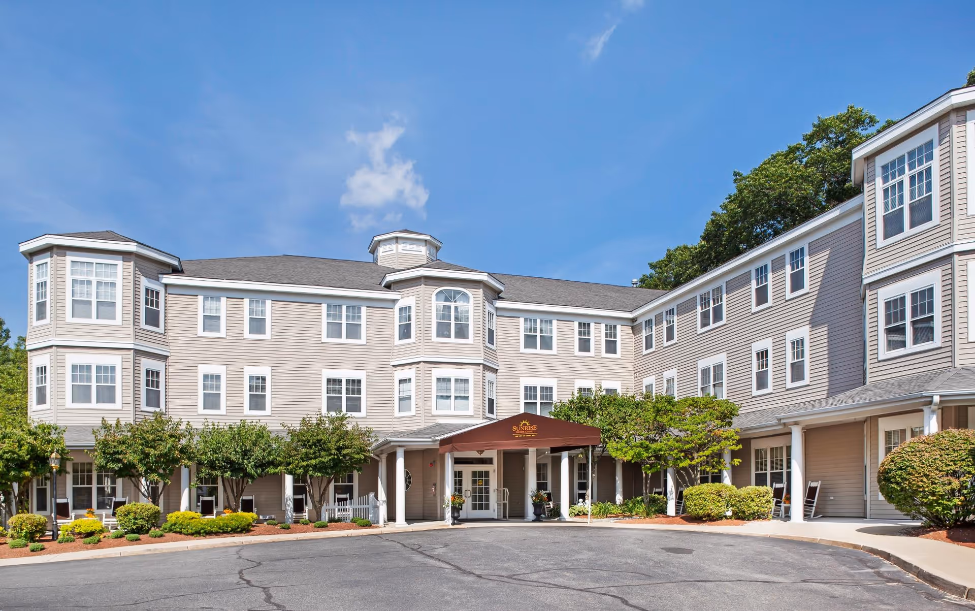 Exterior view of a three-story senior living facility building with beige siding and multiple windows. The entrance has a covered awning with the Sunrise logo and name. There are trees and shrubs around the building and a paved driveway in front.