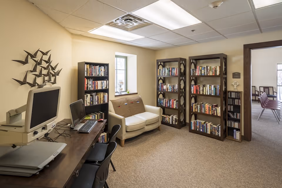 A cozy reading and computer area in a senior living facility featuring two computer workstations with monitors and keyboards on a wooden desk, a small white loveseat with a brown cushion, three tall bookshelves filled with books, a small window letting in natural light, and an adjacent room with chairs visible through an open doorway.