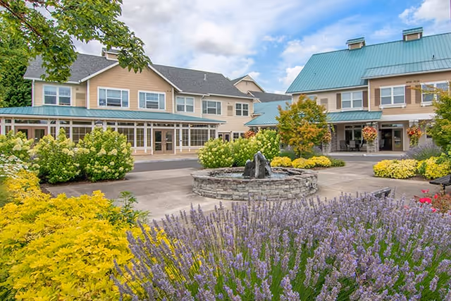 Exterior view of Traditions at Hillside Retirement Community featuring a two-story building with beige siding and a green metal roof. The foreground shows a landscaped garden with purple and yellow flowering plants surrounding a circular stone water fountain under a partly cloudy sky.