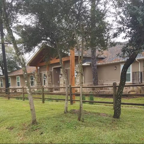 Front exterior of a single-story memory care building with a wooden fence, trees, and a grassy lawn.