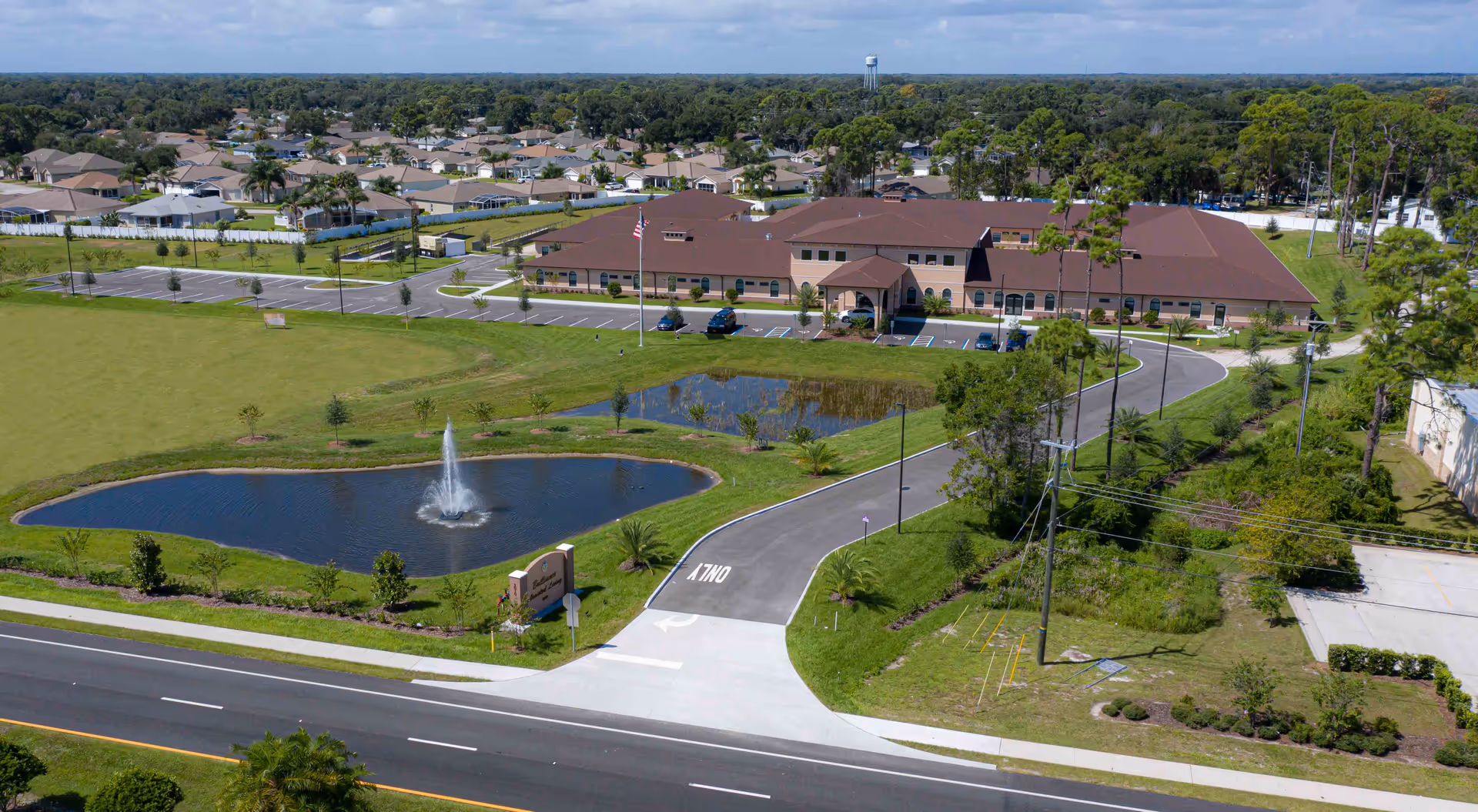 Aerial view of an assisted living building with a curved driveway, parking lot, and a pond with a fountain in front.