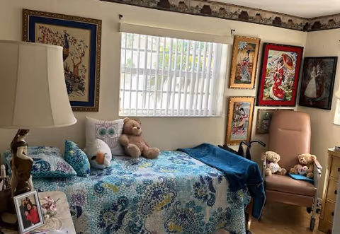 A cozy bedroom with a single bed covered in a blue and green patterned quilt. On the bed are stuffed animals including a teddy bear, an owl pillow, and a duck. A window with vertical blinds is above the bed. The walls are decorated with framed artwork featuring peacocks and other colorful designs. There is a beige armchair with two teddy bears seated on it, and a wooden dresser with a lamp and framed photo on top.