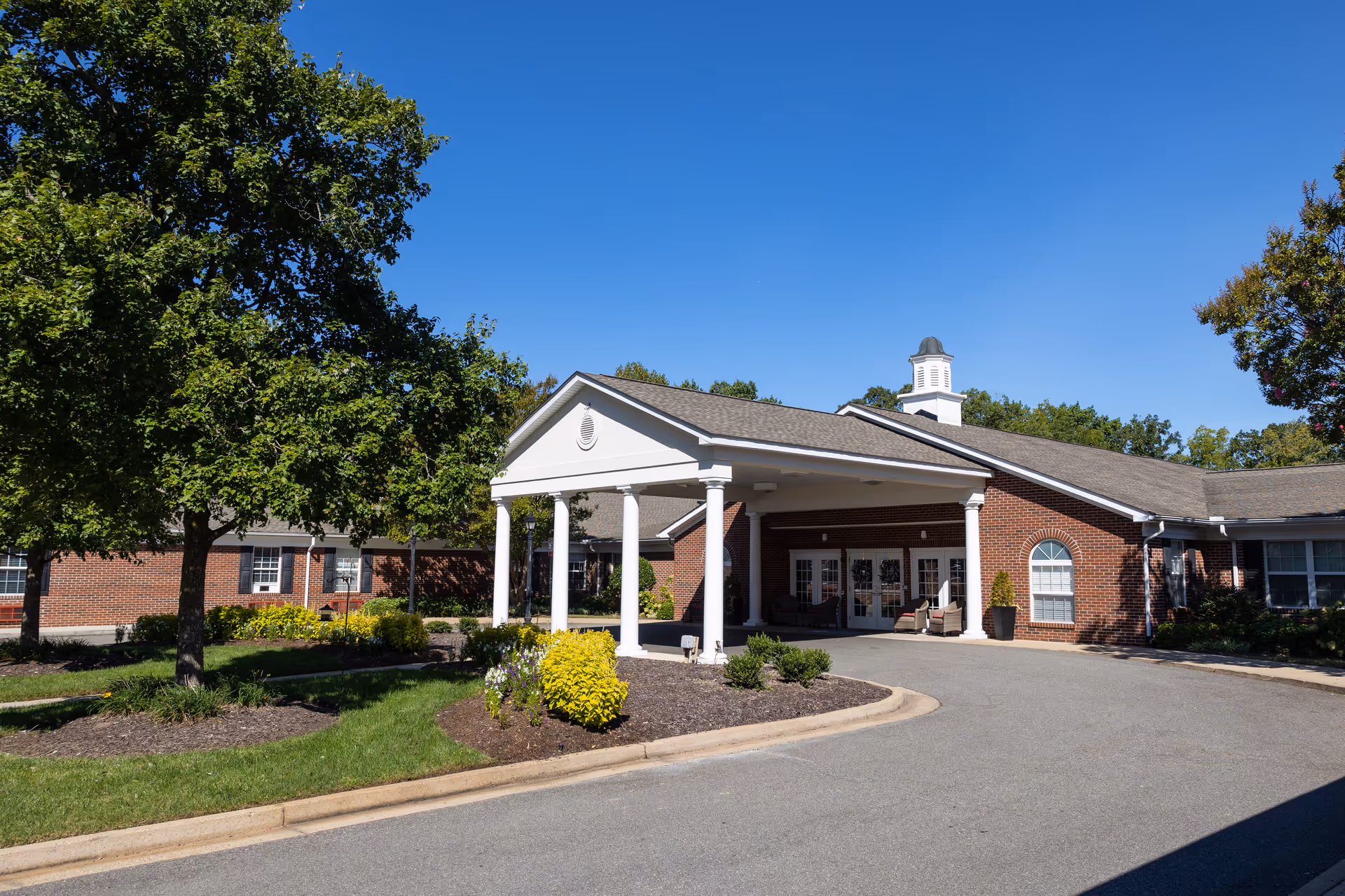 Exterior view of a senior living facility with a covered entrance supported by white columns. The building is made of red brick with white trim and has a cupola on the roof. There are trees, shrubs, and landscaped flower beds surrounding the driveway leading to the entrance under a clear blue sky.