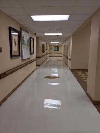 A long, clean hallway in a nursing home or rehabilitation facility with beige walls, handrails on both sides, framed artwork hanging on the left wall, and a shiny tiled floor with a diamond pattern in the center. The ceiling has fluorescent lights.