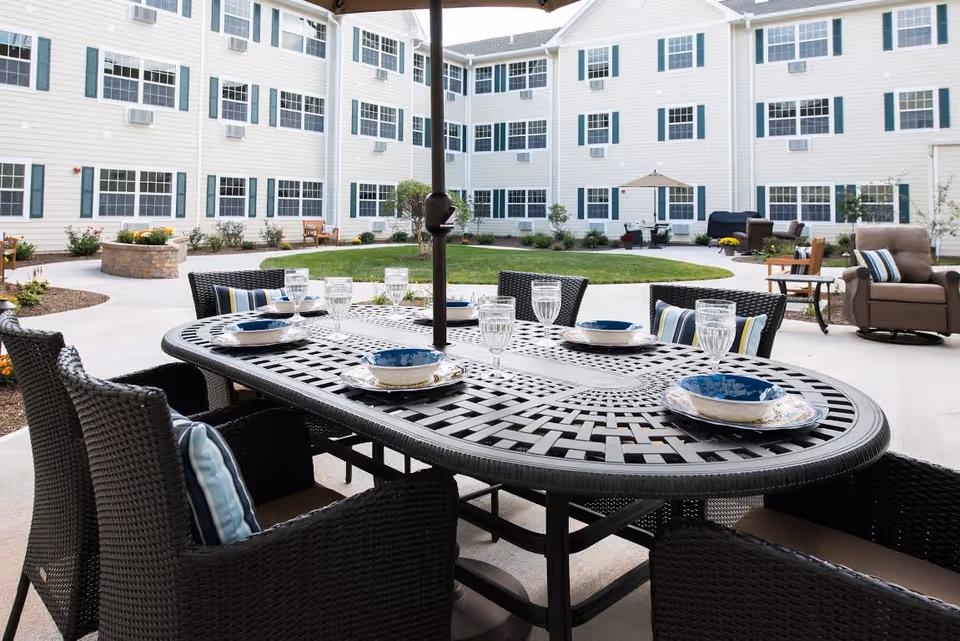 Outdoor courtyard area at The Residence at Glastonbury featuring a black woven patio dining table set with six chairs, each with cushions and pillows. The table is set with plates, bowls, and glasses. In the background, there is a green lawn, a fire pit, additional seating including armchairs and benches, and a three-story building with multiple windows and white siding.